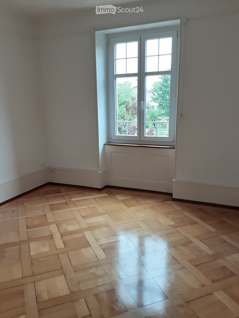 Empty room with wooden parquet flooring, white walls, and a large window with double glass panels. The room has a radiator under the window and is completely unfurnished.