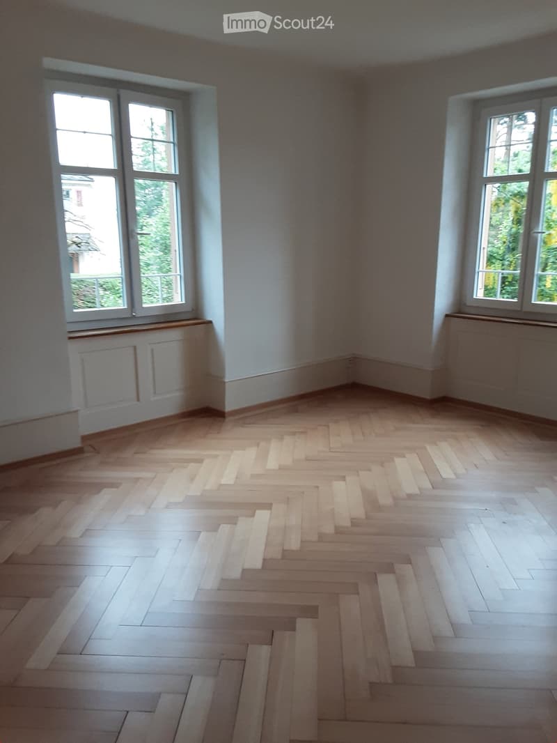 Empty room with light wooden herringbone flooring, white walls, and large windows with white frames. No furniture or equipment present.