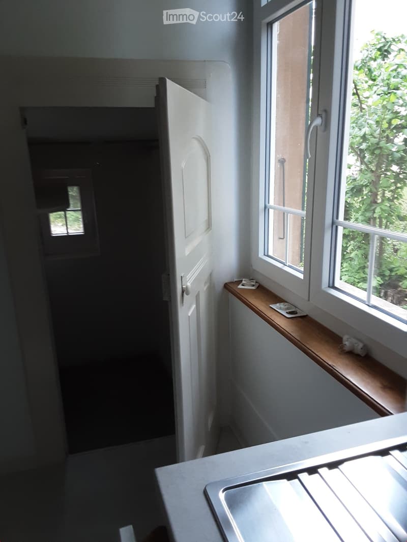 A narrow hallway with white walls, a wooden shelf on the right side, and a white door on the left leading to another room. Natural light streams in from a window on the right.