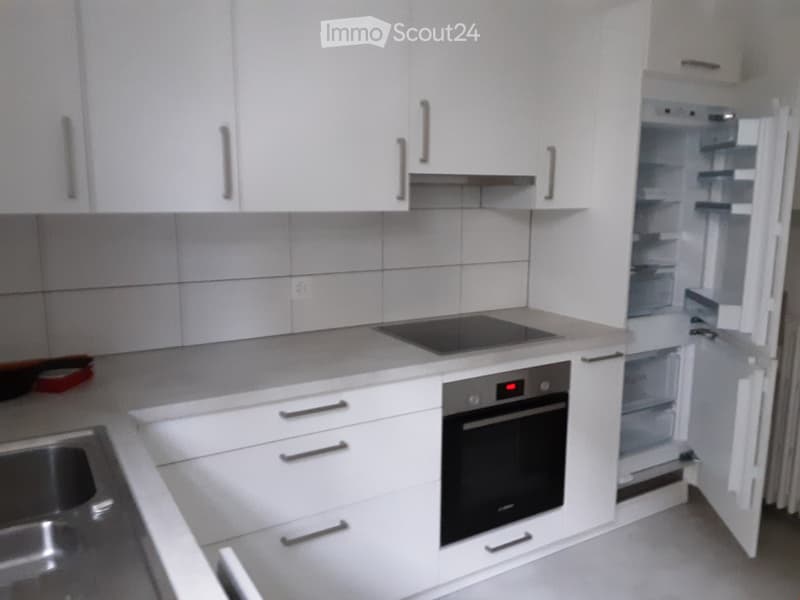 Modern kitchen with white cabinets, stainless steel sink, induction cooktop, oven, and refrigerator. The countertops are light-colored, and the backsplash is tiled.