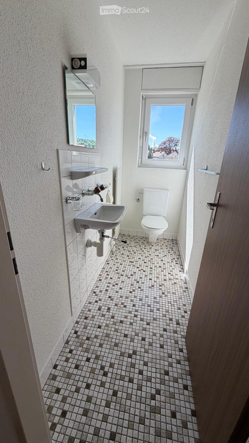 Modern bathroom with white walls, tiled floor, sink, toilet, and mirror. Features a window with a view of the sky and trees.