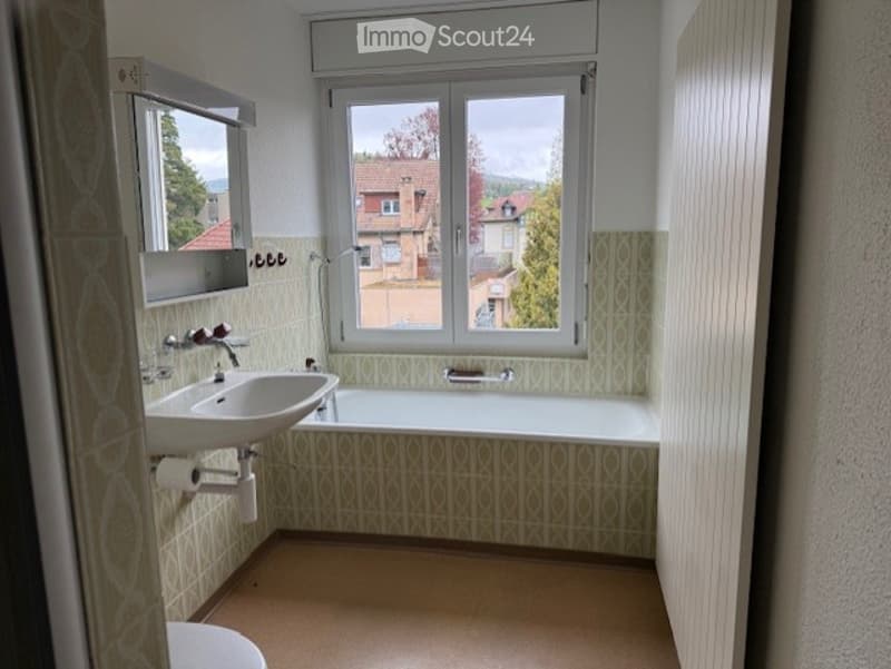 Bathroom with a white sink, bathtub, mirror, and toilet. The walls are tiled with a green and beige pattern, and there is a large window offering a view of the outside.
