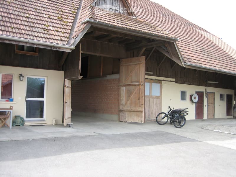A rustic barn with a tiled roof, large wooden doors, and a small attached building with a table and chairs. Two bicycles are parked near the entrance.