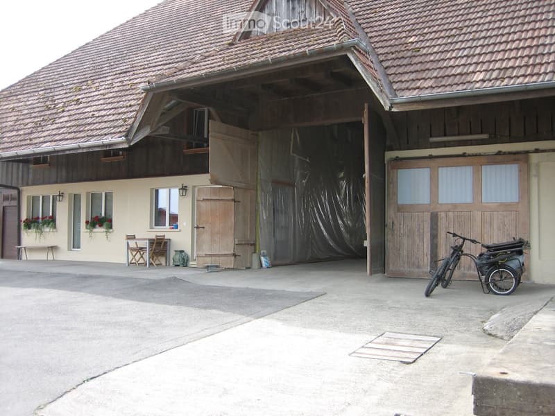 A rustic two-story building with a tiled roof, large wooden doors, and a covered outdoor area. There is a bicycle with a trailer parked near the entrance, and a small table and chairs are visible on the left side.