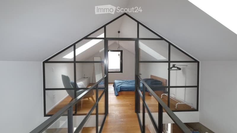 Modern, open-concept attic bedroom with wooden flooring, large windows, and glass partitions. Features a double bed, nightstand, armchair, and clothing rack.
