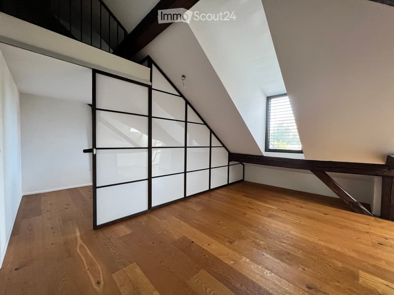 Empty attic room with wooden flooring, white walls, and a large window with blinds. The room features a modern sliding door with a geometric design.