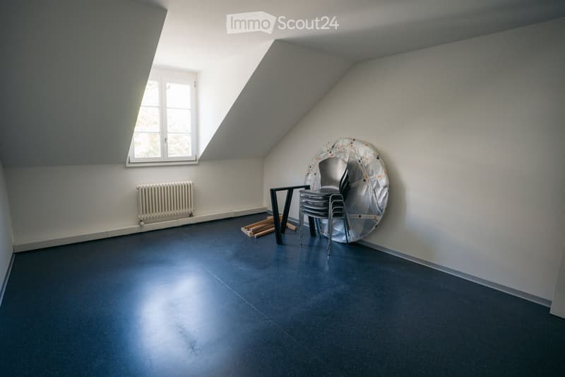 An attic room with a slanted ceiling, dark blue floor, white walls, and a window. Contains a small table, a stack of chairs, and a radiator.
