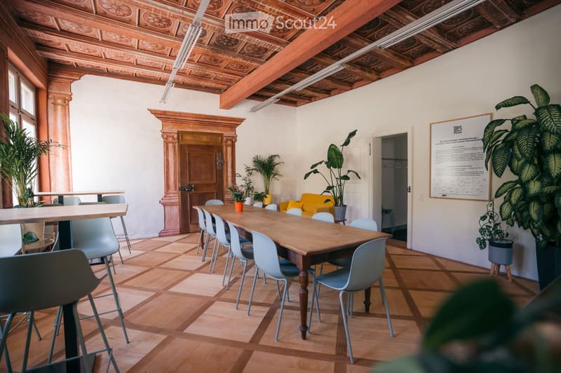 Bright, modern room with wooden flooring, a long wooden table surrounded by chairs, a yellow sofa, and several potted plants. The room features exposed wooden beams and a decorative ceiling.