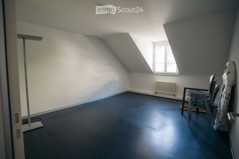 An attic room with slanted ceilings, blue flooring, and white walls. A window with white frames allows natural light to enter. A radiator is mounted on the wall, and several chairs are stacked against the wall.