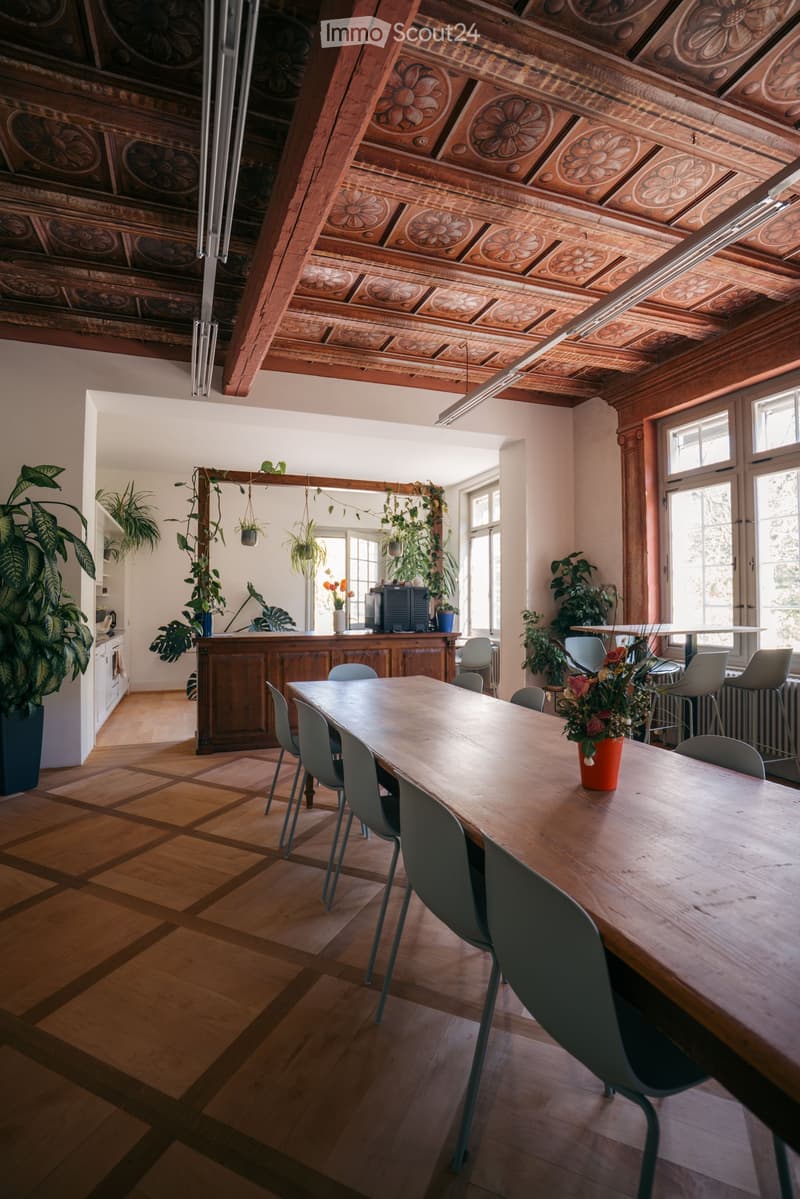 A spacious room with wooden floors and a high, intricately designed wooden ceiling. Features include a long wooden table surrounded by chairs, a bar area with stools, potted plants, and a large mirror on the wall.