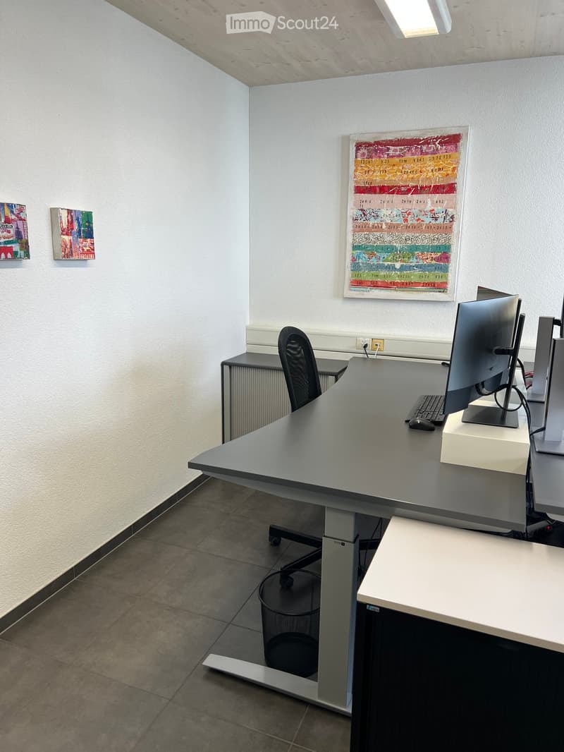 Office space with a large desk, ergonomic chair, computer monitor, keyboard, mouse, and a trash can. The room features white walls, tiled flooring, and colorful artwork on the walls.