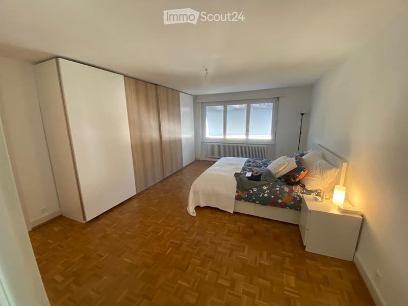 Bedroom with wooden flooring, large window, bed with colorful bedding, nightstand with lamp, and built-in wardrobe.