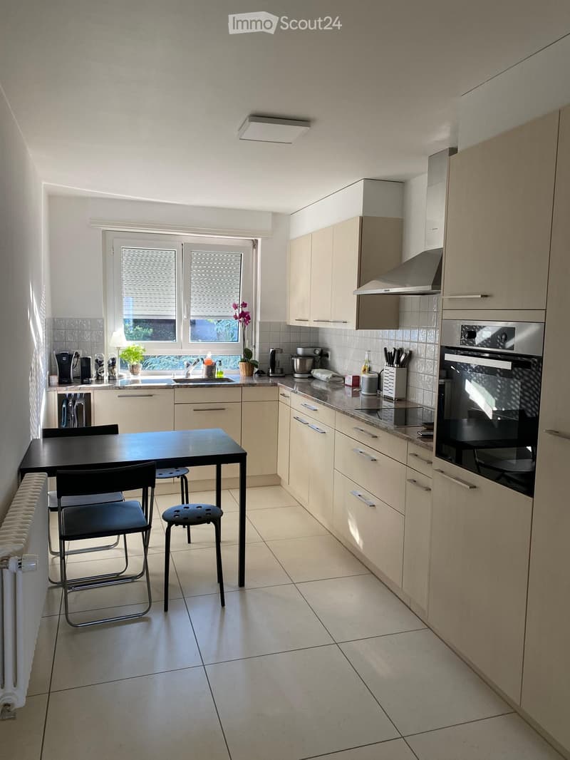 Modern kitchen with light-colored cabinets, a black dining table with chairs, and various appliances including an oven and fridge. The space features a window with blinds and a radiator along the wall.