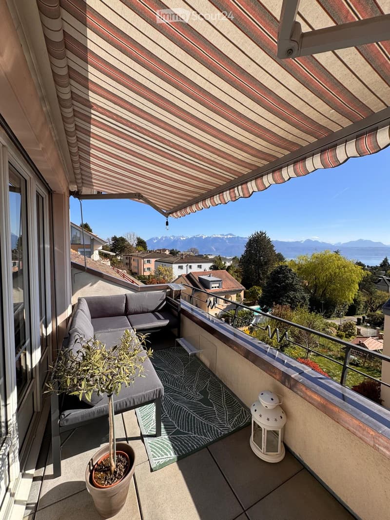 Balcony with a grey sectional sofa, potted plant, decorative rug, and lantern, overlooking a scenic mountain view.