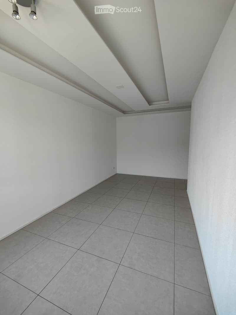 Empty room with white walls, grey tiled floor, and recessed ceiling lighting.
