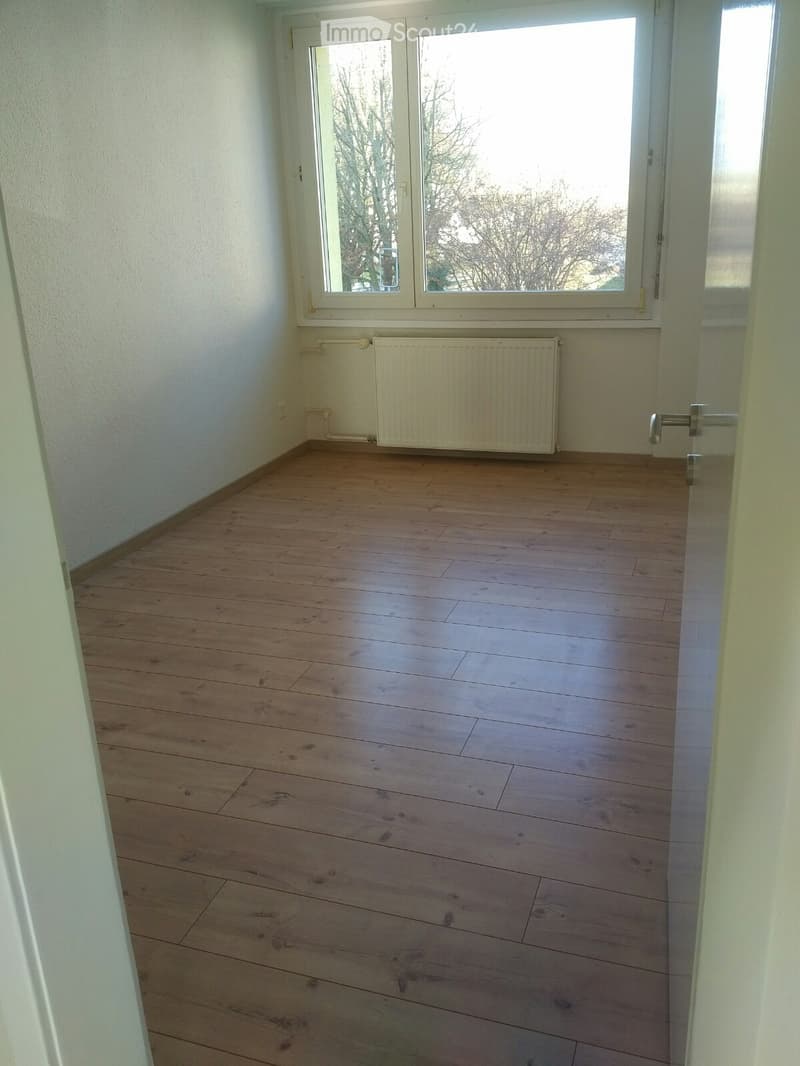 An empty room with wooden floor and white walls, there are windows at the back wall and a white radiator.