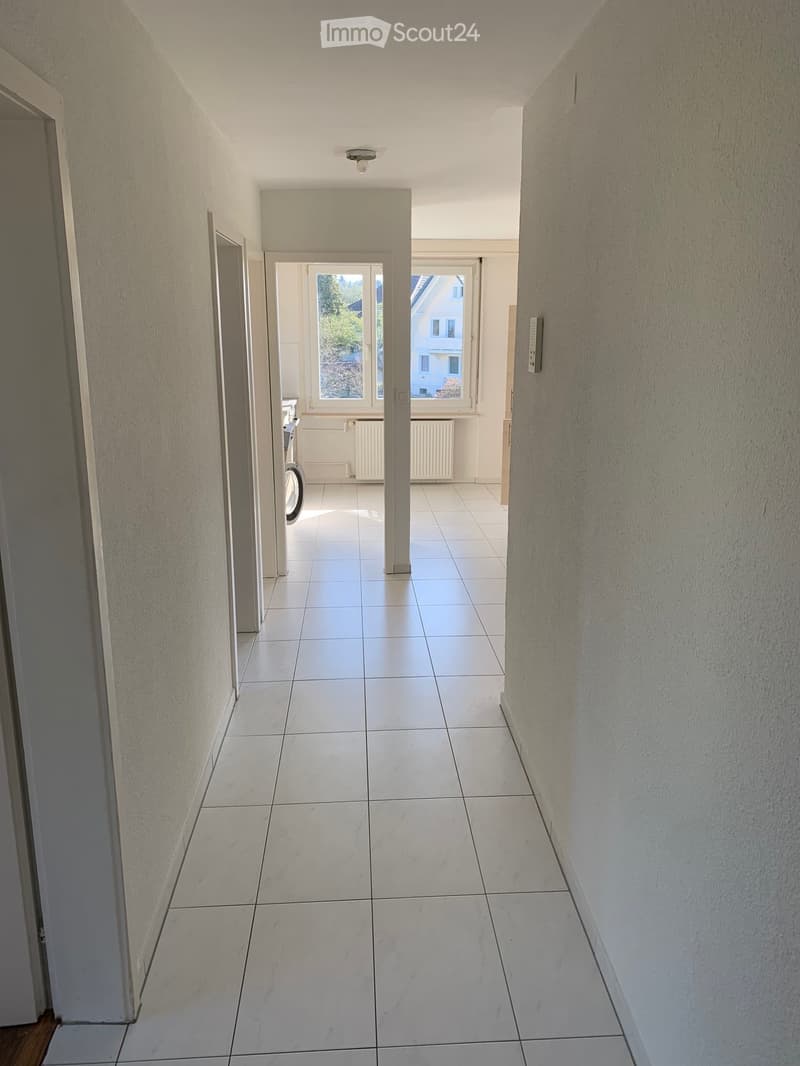 A hallway with white tiled floors and walls, an open room with white windows and white radiators, and an open door leading to an outdoor area with trees and a house.