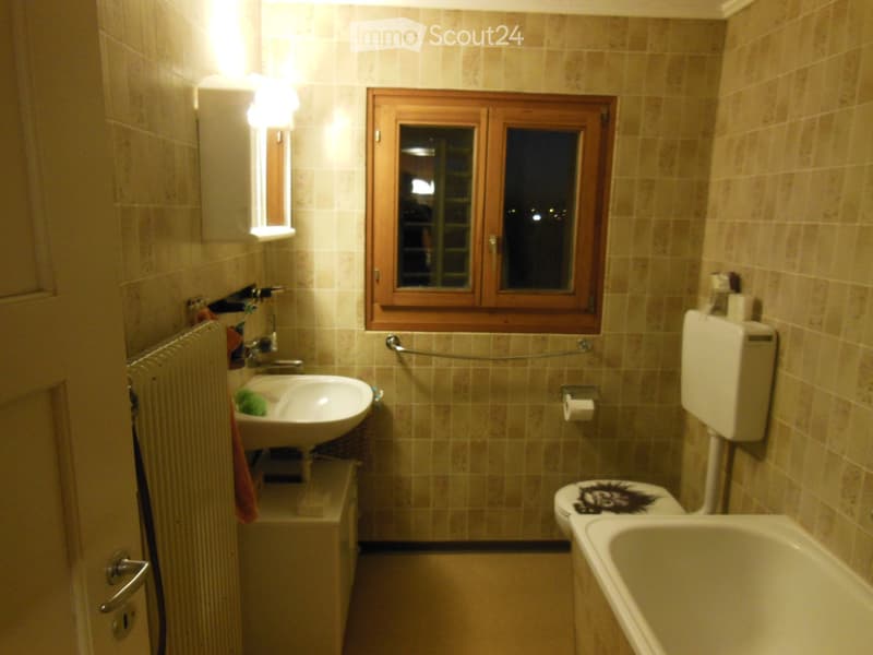 Bathroom with beige tiled walls and floor, featuring a sink, toilet, bathtub, mirror, and towel. A window with wooden frame is visible above the sink.