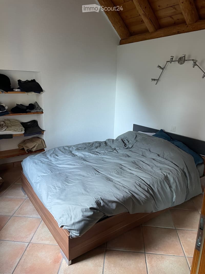 Cozy bedroom with a wooden bed, grey bedding, built-in wooden shelves holding folded clothes and bags, and a modern wall-mounted lamp.