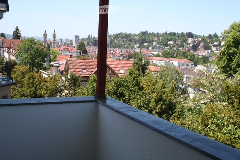 Balcony with a panoramic city view, featuring red-tiled roofs, trees, and distant spires.