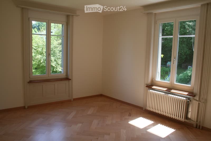 Empty room with wooden flooring, large windows, radiators, and natural light. No furniture or furnishings present.