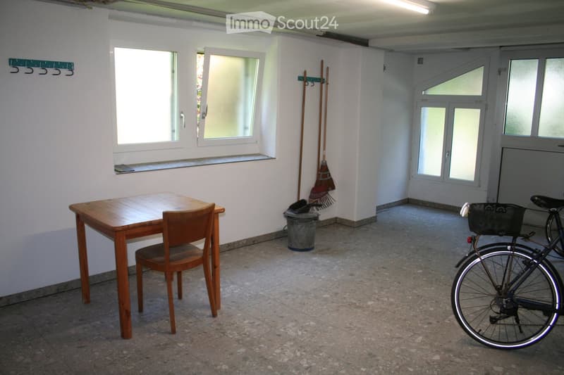 A small, unfurnished room with a wooden table and chair, a bicycle, and cleaning tools near the door.