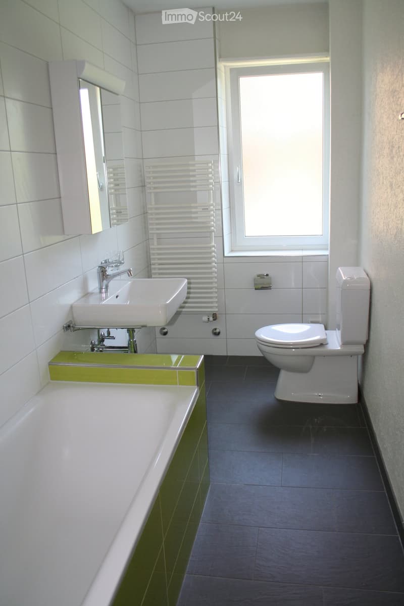 Modern bathroom with white tiles, a white sink, a bathtub, a toilet, a radiator, and a mirror above the sink. The floor is dark grey tiles, and the walls are white with green accents.