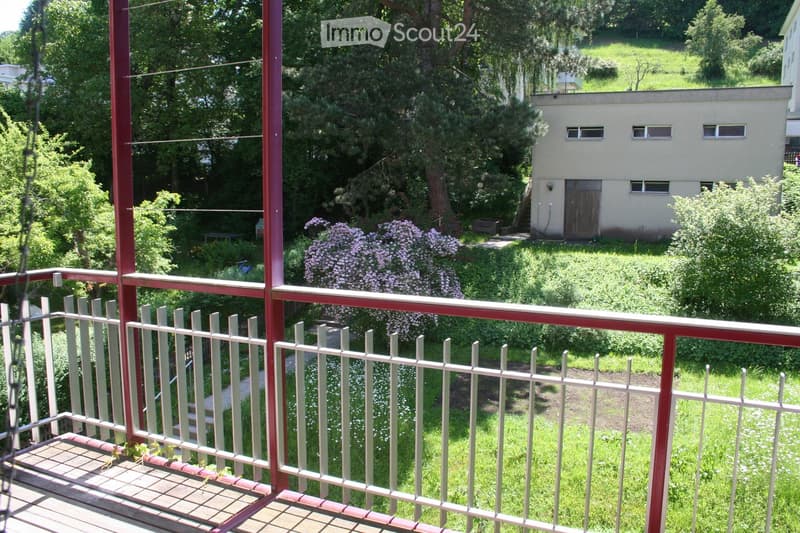 Balcony with wooden decking, metal railing, and view of a garden with flowering bushes and a small building.