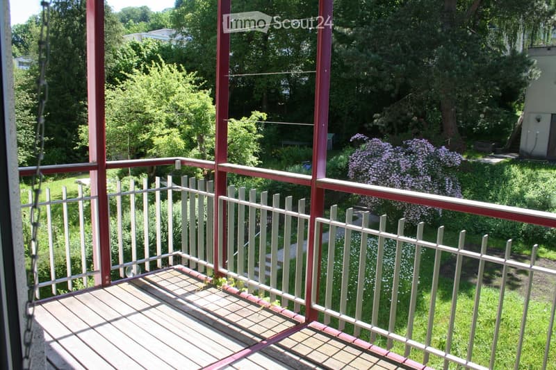 Balcony with wooden decking, white metal railing, and red metal posts, overlooking a green garden with flowering shrubs and trees.