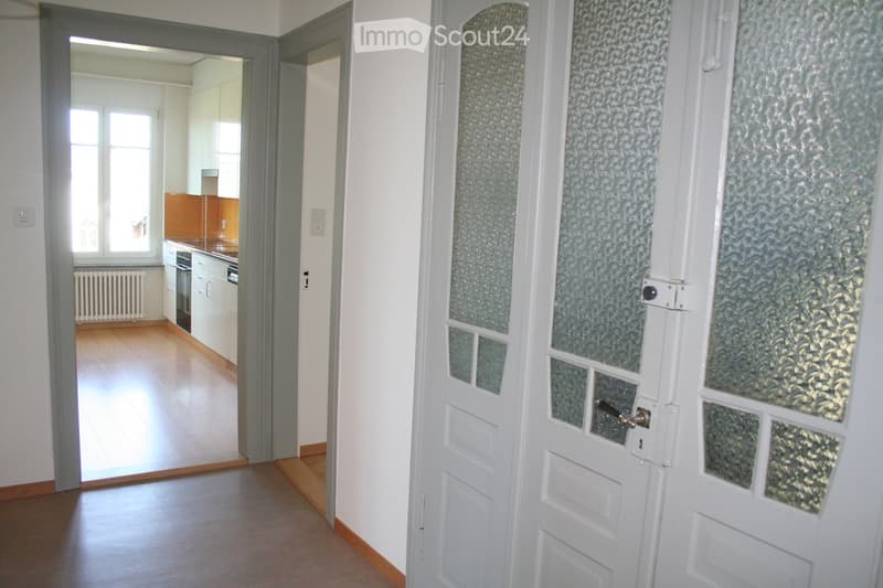 A hallway with a white door featuring frosted glass panels leading to a kitchen area with white cabinets, a countertop, and a window.
