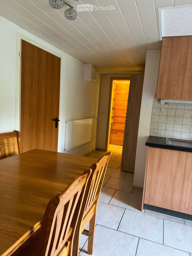 Dining area with wooden table, chairs, and cabinets, leading to a sauna room with wooden walls and a glowing light.