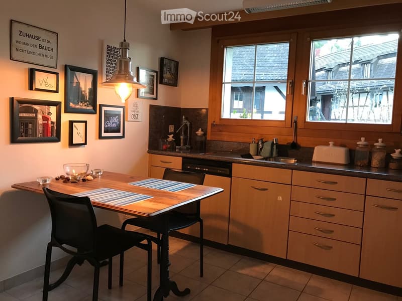 Bright kitchen with wooden cabinets, a central island table, and a sink area. The space features framed artwork and signs on the walls, along with a hanging pendant light.