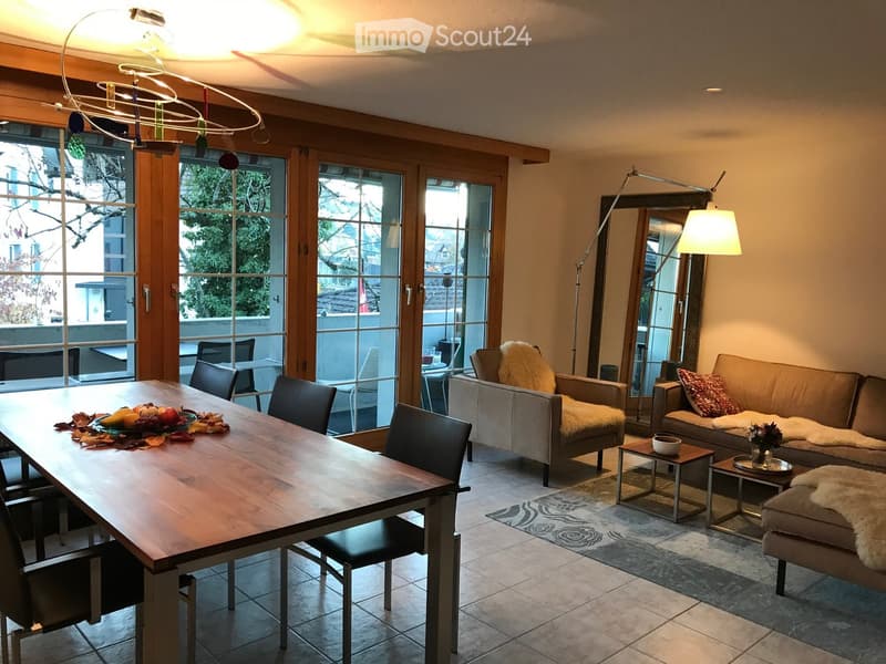A modern living and dining area with a large wooden dining table, black chairs, a grey sofa with a fur throw, a side table, and a floor lamp. The space features large windows with wooden frames and a decorative rug on the floor.