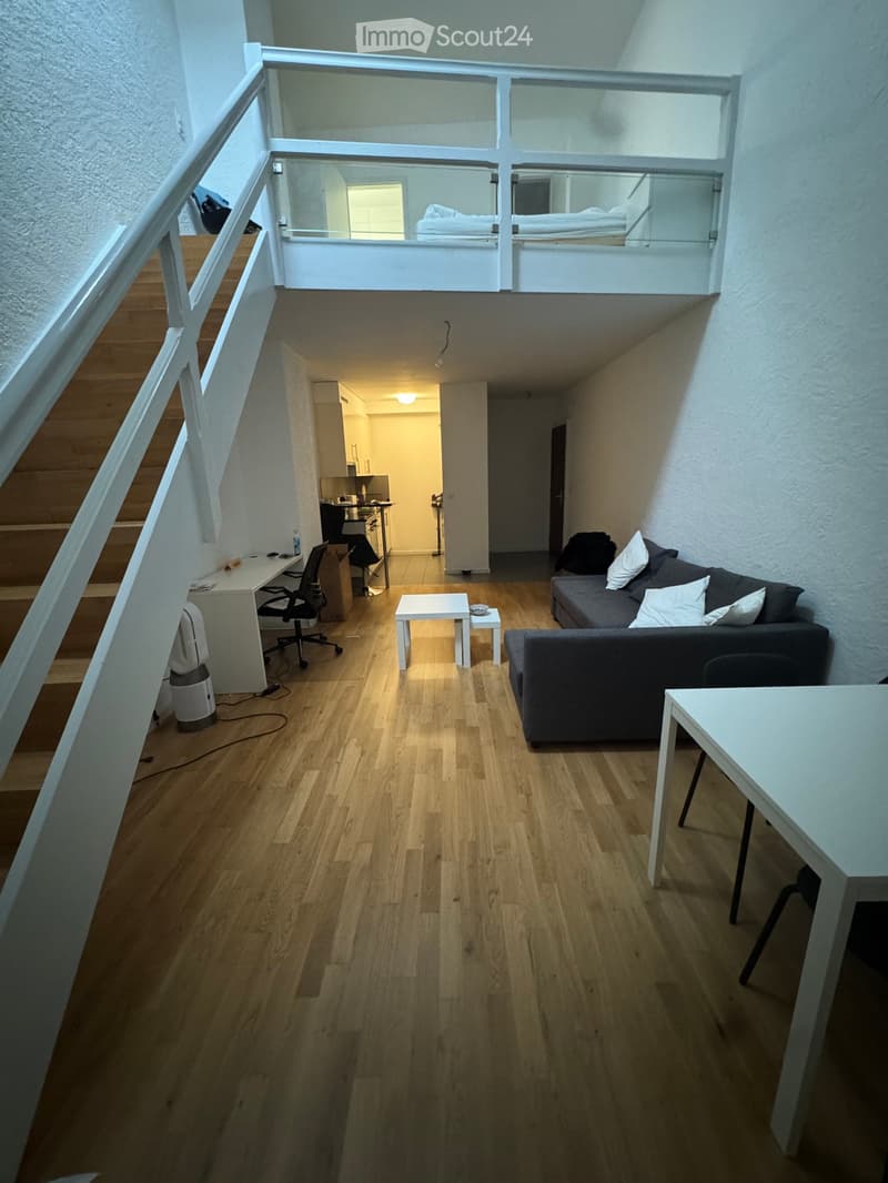 Modern, multi-level living space with wooden flooring, a sofa, small table, desk, and a lofted bedroom area with a bed.