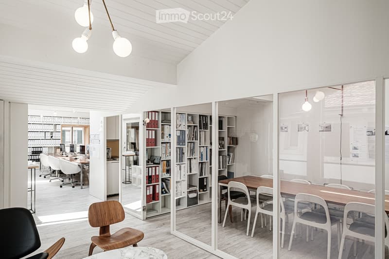 Bright, modern office space with white walls, wooden flooring, and large glass partitions. Features include a mix of wooden and white chairs, tables, shelves filled with books and binders, and pendant lighting.