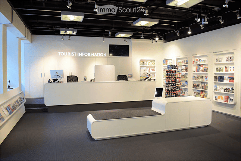 Tourist information center with a white reception desk, two chairs, a screen, shelves with brochures, and a bench.