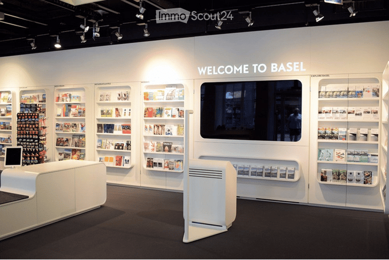 Modern, minimalist room with white walls and black carpeting. Features a white sofa, a large screen mounted on the wall, multiple shelves filled with books and decorative items, and a small table with a laptop. The room is well-lit with track lighting.