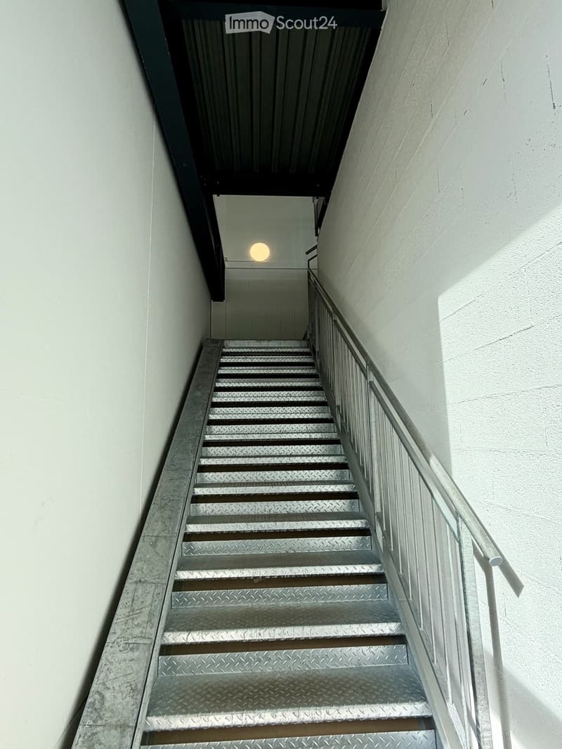 Metal staircase with a handrail, leading upwards, with a single light fixture visible at the top.