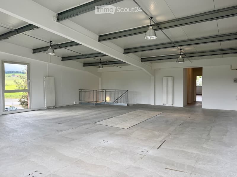Empty room with white walls, concrete floor, large windows, metal staircase, and industrial-style lighting fixtures.