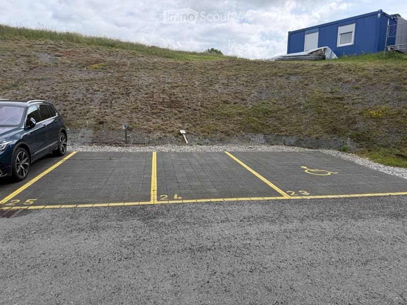 Parking lot with marked spaces, a dark-colored SUV parked in one of the spaces, and a blue container in the background on a grassy slope.