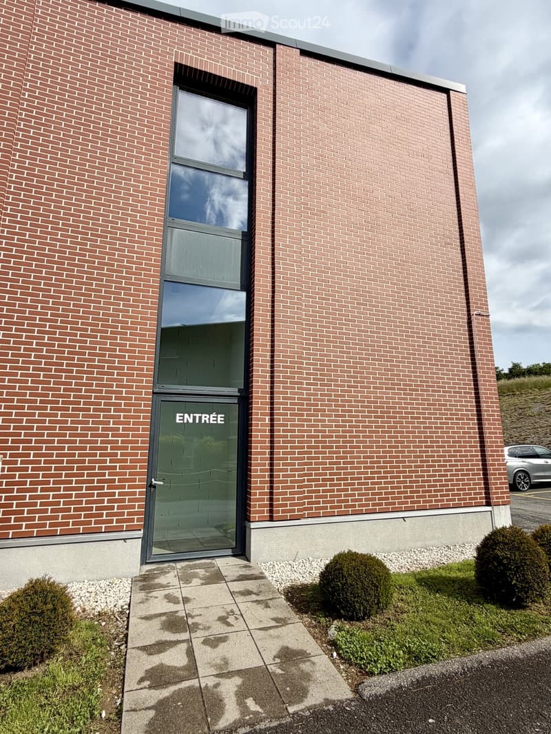 Modern brick building entrance with glass door labeled "ENTRÉE" and small landscaped area.