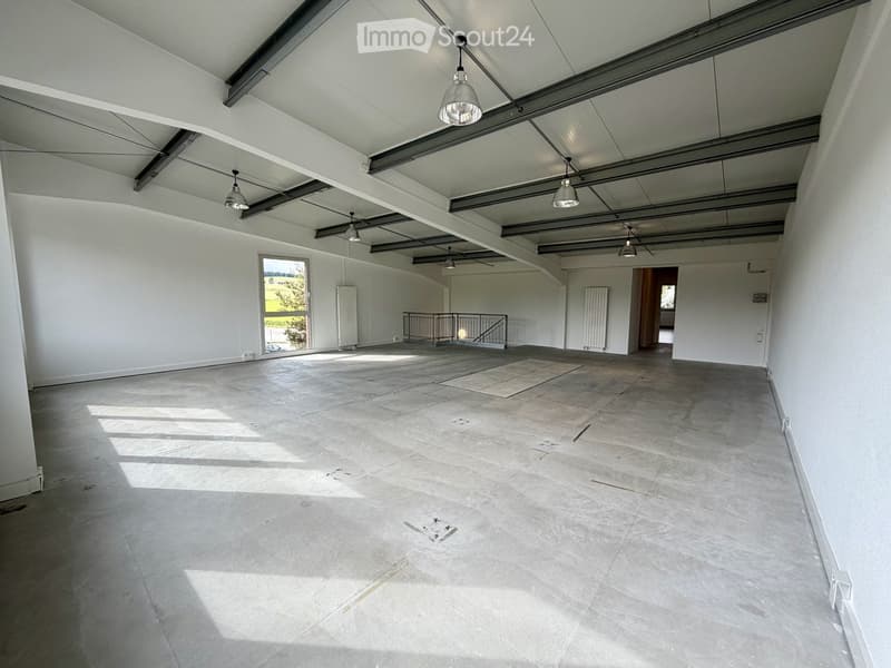 Empty room with white walls, concrete floor, exposed metal beams, and pendant lights. Features a large window and a staircase leading to another level.
