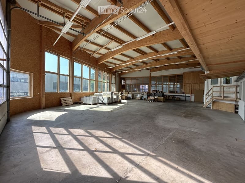 Large, open, unfurnished space with wooden walls and ceiling, concrete floor, large windows, and some stacked materials and storage items in the background.