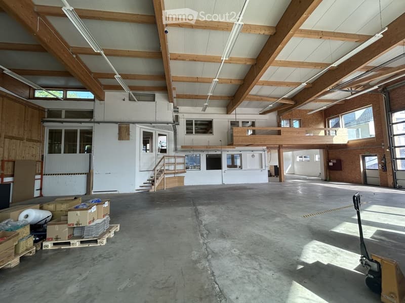 Large, open industrial-style space with exposed wooden beams, concrete floor, and minimal furnishings. Features include a staircase leading to an upper level, large windows, and some stacked boxes and pallets in the foreground.