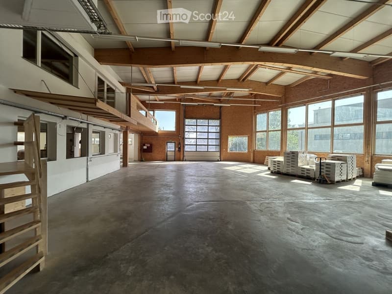 Large, open, unfurnished space with exposed wooden beams, concrete floor, and large windows allowing natural light. Stacks of materials and a pallet jack are visible, indicating a storage or workshop area.