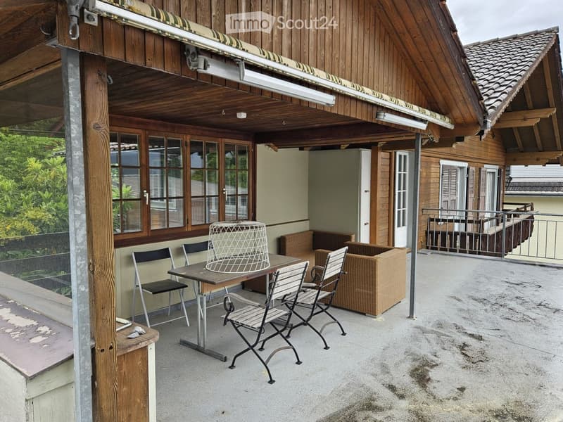 Balcony with wooden table, chairs, and a wicker sofa, overlooking a garden area.