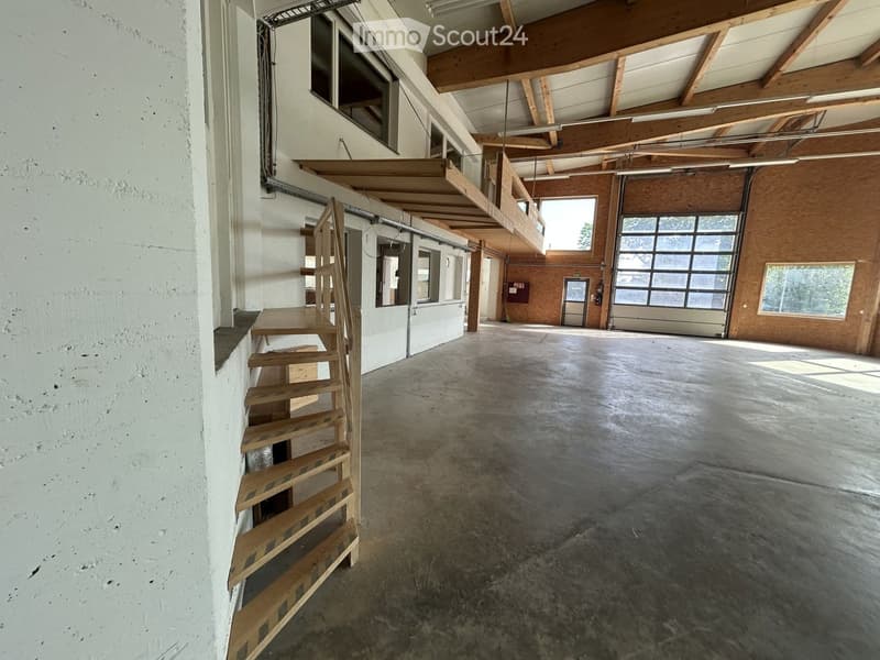 Empty industrial-style space with concrete floor, exposed wooden beams, large windows, and a wooden staircase leading to an upper level.