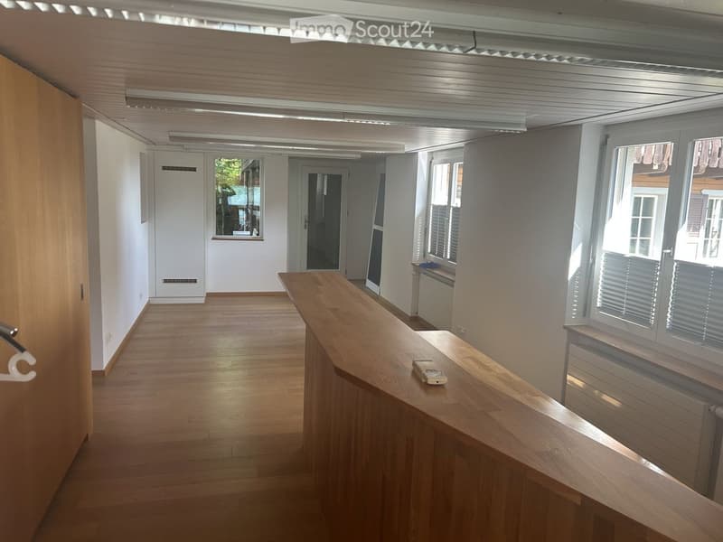 Empty room with wooden flooring, white walls, and large windows. A wooden reception desk is present, but no furniture or personal items are visible.