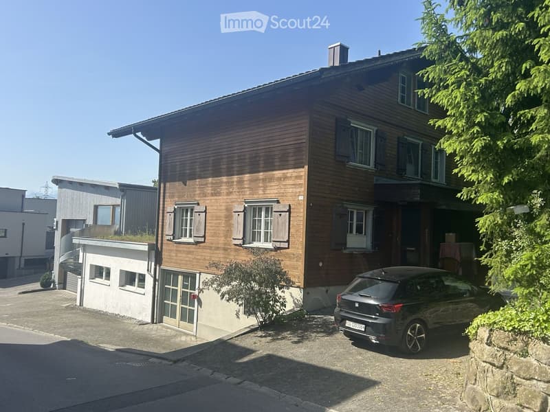 Two-story wooden house with multiple windows, shutters, and a small balcony. A car is parked in front, and there is a small garden area with plants and a stone wall.