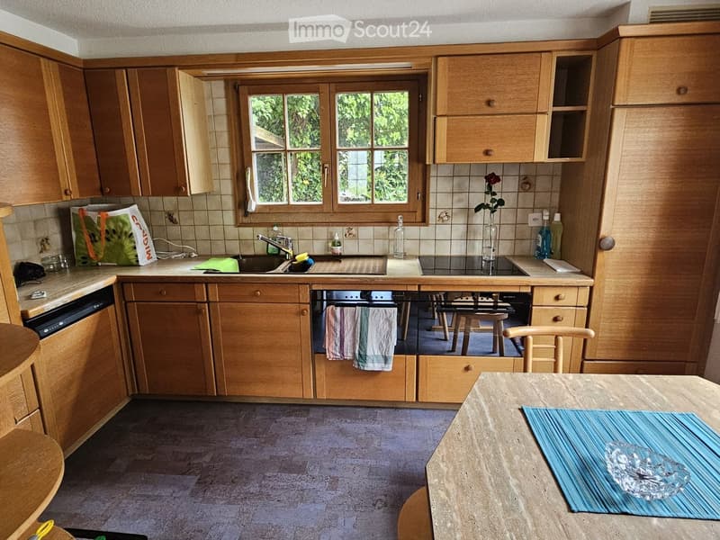 A cozy kitchen with wooden cabinets, a sink, an oven, a dishwasher, and a dining table with a chair. The space features a window with a view of greenery, and various items such as a handbag, bottles, and a towel are visible.